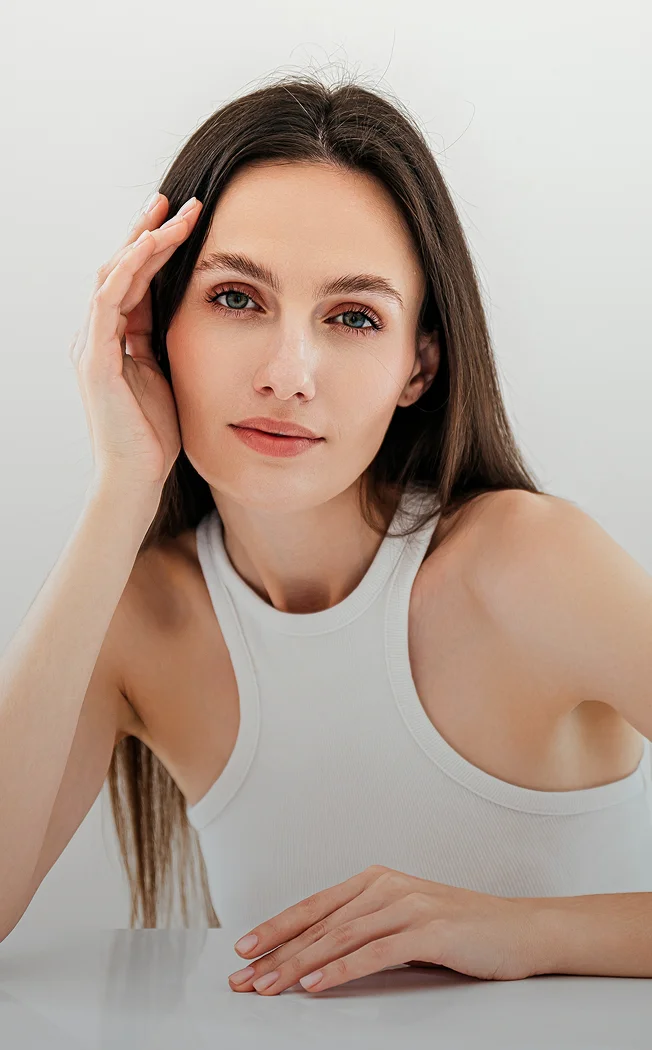 Young woman with long brown hair in white tank top posing with hand near face against neutral background - eyes in New York , NY