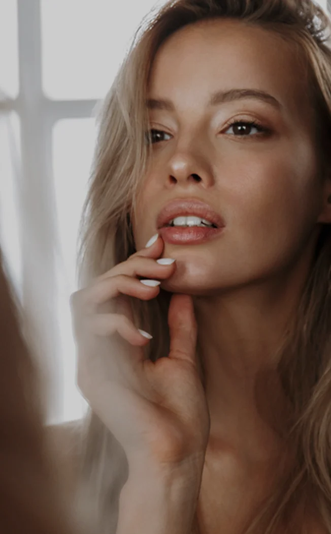 Beautiful blonde woman with natural makeup touching face in soft natural lighting near window - in New York , NY