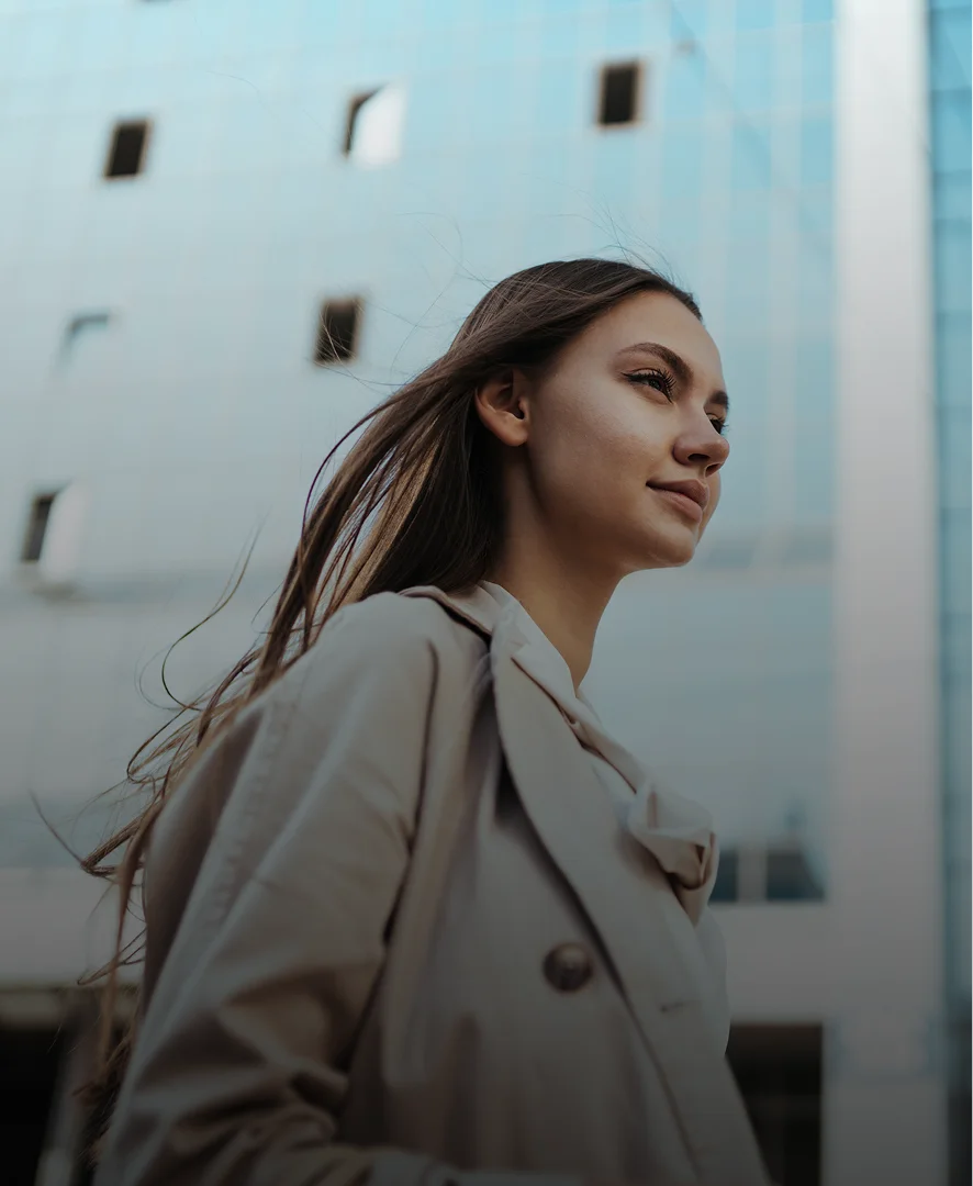 Young woman in beige trench coat with windblown hair against urban sky backdrop - in New York , NY