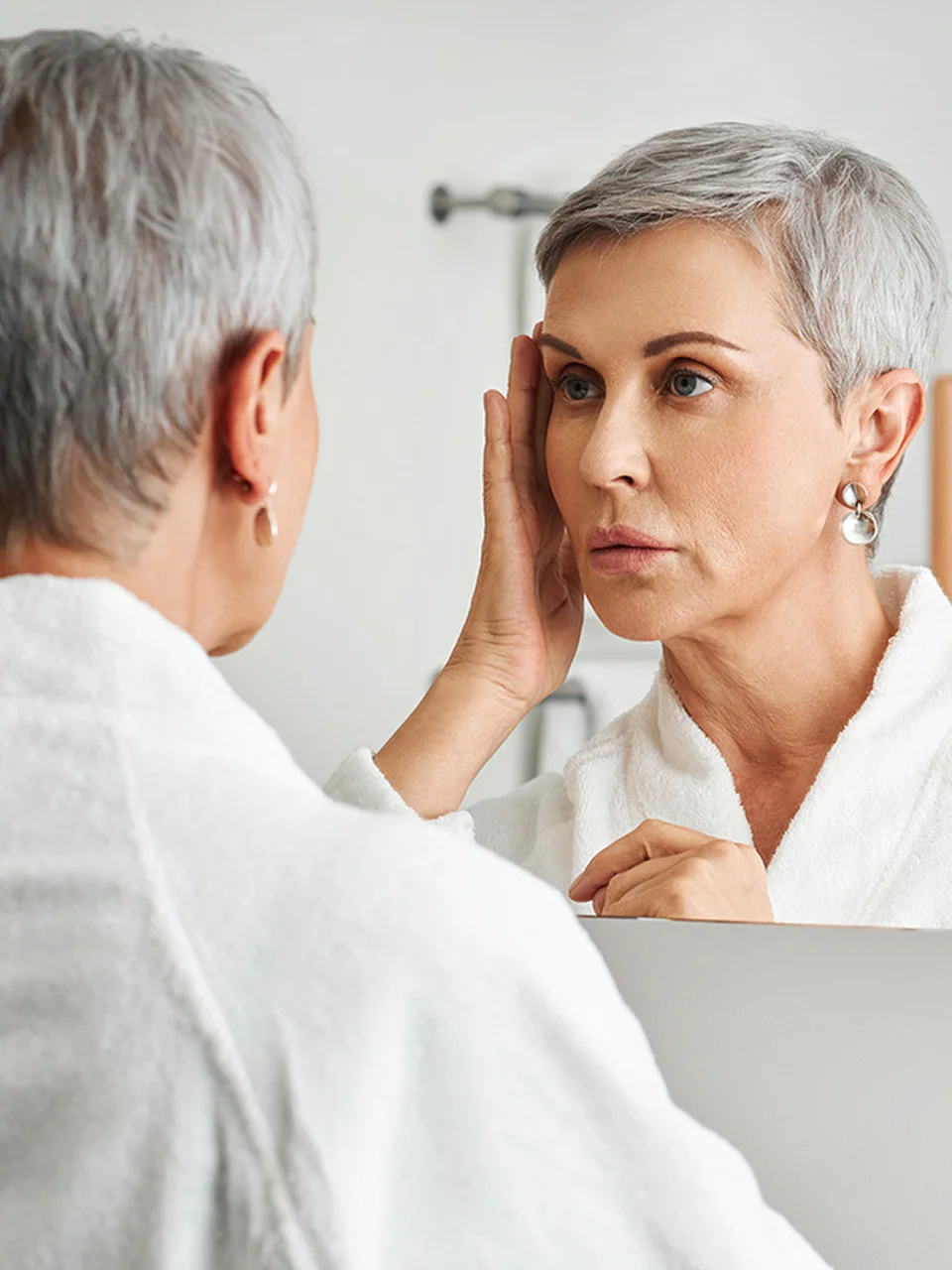 Woman with short gray hair examining her face in bathroom mirror for skincare and aging concerns - in New York , NY