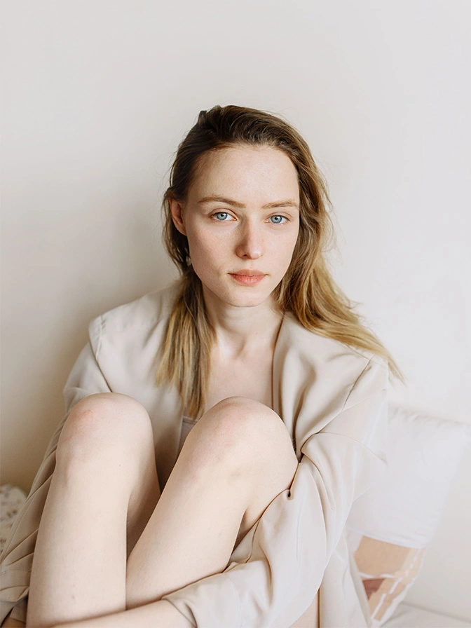 Young woman with long blonde hair in white clothing sitting against neutral wall in natural light - lower eyelid lift in New York , NY