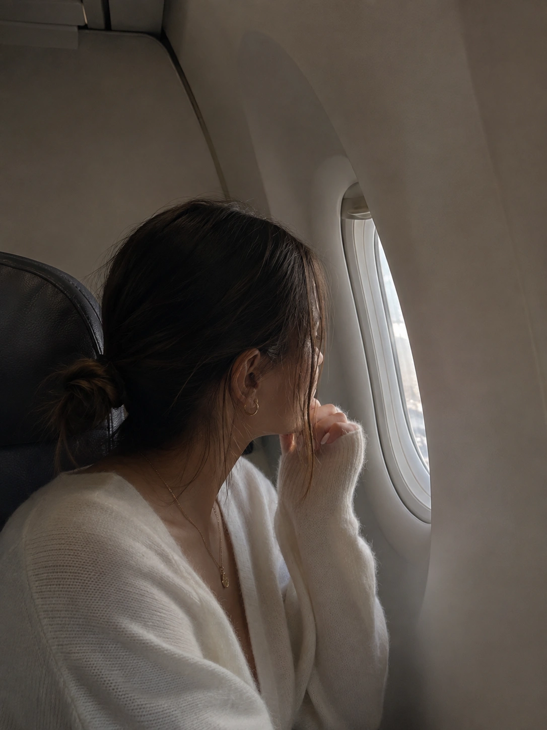 Woman looking out airplane window during flight, wearing white clothing - in New York, NY